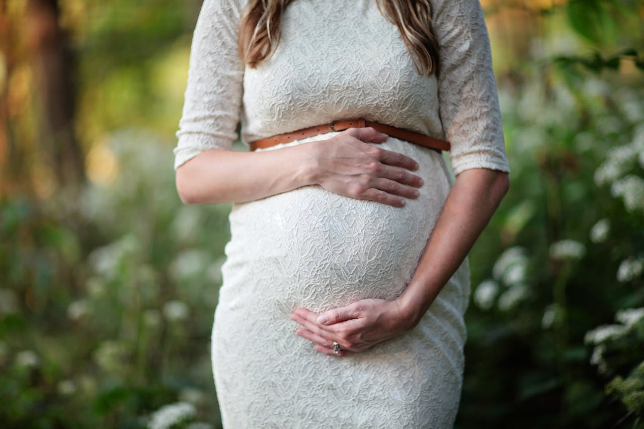 about-us-02 A pregnant woman in a lace dress gently cradling her belly in an outdoor setting.