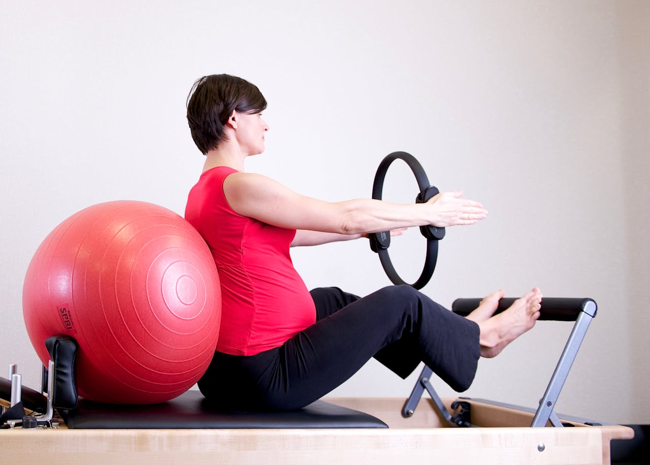 about-us-01 A pregnant woman in a red top practicing Pilates using a fitness ring and exercise ball in a gym setting.